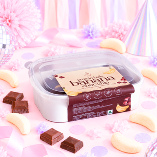 A tub of Protein Labs banana chocolate chip protein balls sits on a pink party-themed surface with scattered chocolate chips and banana pieces. The clear-lidded container shows the product inside