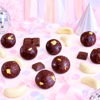 Banana Lolly Chocolate protein balls and chocolate bars arranged on a pink party-themed surface with confetti, banana lollies, a disco ball, and a party hat.