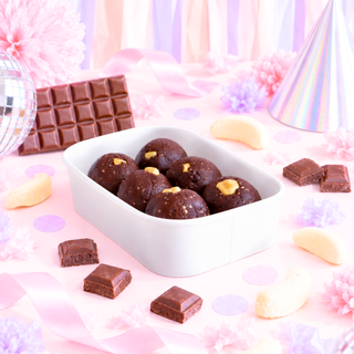 A white rectangular dish filled with banana lolly protein balls, styled on a pink party surface with scattered chocolate squares, banana lollies, a chocolate bar, a disco ball, and a party hat.