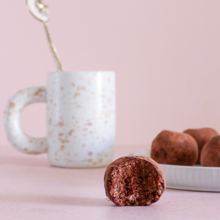Half bitten choc teaser protein ball resting near a plate of them with a mug full of hot chocolate