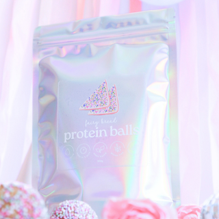A holographic silver resealable pouch labeled 'fairy bread protein balls' with a pink sprinkle-sprinkled triangle logo, photographed against a soft pastel pink and white draped background with blurred protein balls at the bottom.