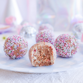 A close-up of fairy bread protein balls coated in sprinkles, with one ball broken in half in the foreground to show the crumb texture. The balls are arranged on a white plate with a blurred festive background.