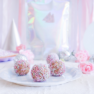 Four fairy bread protein balls topped with pink, white, and green sprinkles arranged on a white paper plate, with a blurred iridescent protein ball mix pouch and pink decorative flowers in the background.