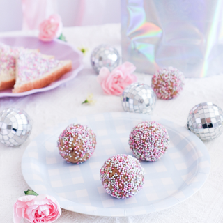 Four fairy bread protein balls topped with pink, white, and silver sprinkles are arranged on a light blue and white checkered paper plate. The scene is decorated with pink flowers, mini disco balls, and a slices of fairy bread on a lavender plate in the background, all under soft, bright lighting.