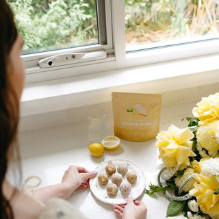 Person preparing food on a kitchen counter with lemons, lemon cheesecake protein balls and flowers.
