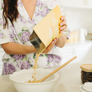 Woman in a kitchen pouring ingredients into a bowl with a wooden spoon.