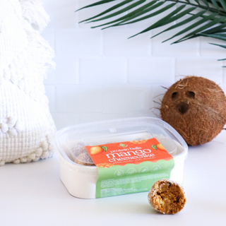 Container of mango cheesecake flavoured protein balls with a coconut on a white surface