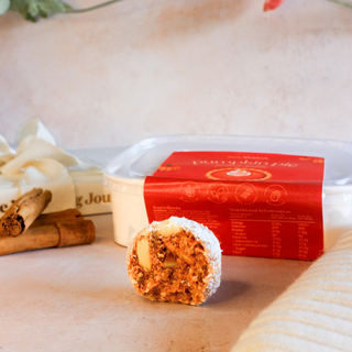 A pumpkin pie flavored protein ball, bitten open to show its textured filling, placed in front of its white and red packaging container on a beige surface.