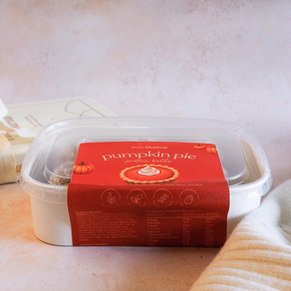 A white plastic tub of pumpkin pie protein balls with a clear lid and a rusty orange paper label reading 'pumpkin pie' set on a soft pink surface with a folded cream fabric beside it.