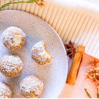 Five Pumpkin Pie Protein balls dusted with coconut, with one bitten piece showing inside arranged on a speckled white plate. The plate sits on a cream ribbed textile alongside cinnamon sticks, star anise, and small orange flowers.