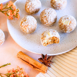 Several round, Pumpkin Pie Protein Balls arranged on a speckled white plate, with one ball broken open to show the textures inside. The scene includes cinnamon sticks, star anise, small orange flowers, and a light wood surface in a styled food presentation.