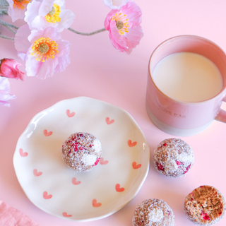 Pink mug with milk, a plate with heart patterns, and raspberry white chocolate protein balls on a pink background with flowers.