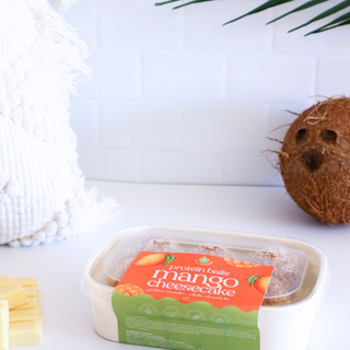 Protein ball container with 'mango cheesecake' flavour label on a white surface with a coconut and leaf in the background.