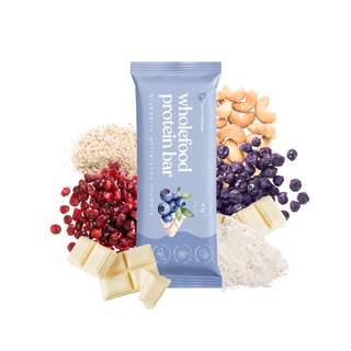 A Blueberry White Choc Crumble wholefood protein bar in a light blue wrapper, surrounded by its ingredients: white chocolate chunks, cranberries, blueberries, cashews, and pea protein, on a solid white background.
