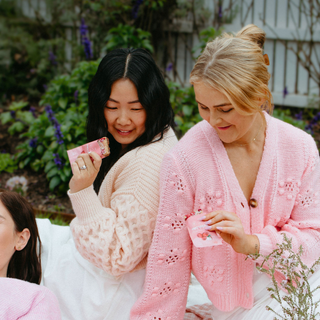 Three women wearing pink cardigans sitting outdoors with plants in the background