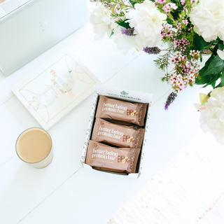 A box of Hazelnut Choc Fudge protein bars lay on a white desk with a wellness book, coffee and fresh flowers 