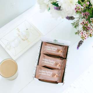 Three brown protein bars in a white box labeled 'better being protein bar' on a white tabletop, styled with flowers and a small cup of coffee.