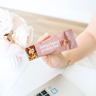 A hand holding a protein bar wrapper labeled 'better being protein bar' over a white desk with a laptop and pale pink flowers in the background.