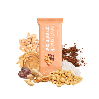 A package of a peanut butter snick snack protein bar stands upright on a white background, surrounded by peanuts, a swirl of peanut butter, chocolate pieces, and cocoa powder. The package is orange-colored with white text reading 'snick snack protein bar' and 'peanut butter chocolate flavour'.