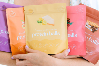 5 packets of protein ball mixes being held in the arms of a woman 