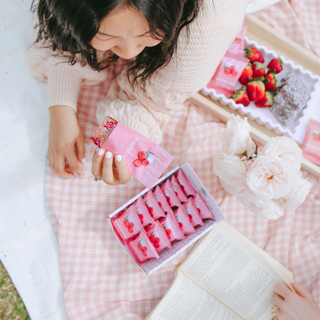 A woman in a cream knit sweater sits on a pink-and-white checkered blanket outdoors, holding a pink packet of strawberries. An open box filled with raspberry lamington nourish bites lies nearby, with a tray of fresh strawberries and a book on the blanket.