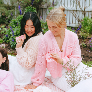 Two women sitting outdoors on a pink-and-white checkered blanket, wearing light pink knit cardigans and white tops. Holding Sneaky Wholefoods Raspberry Lamington Nourish Bites 