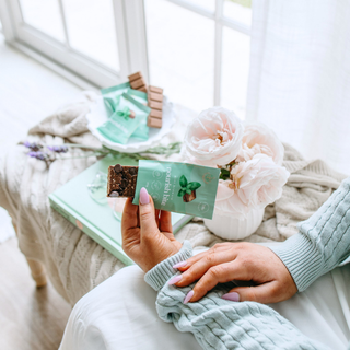 A person holds a mint green nourish bite, with more  arranged on a light-colored bench in a bright room with soft pink flowers.
