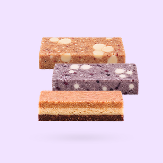 Three protein bars stacked vertically on a white background. The top treat is light brown apple cinnamon with white chocolate cream spots, the middle treat is purple blueberry with white chocolate spots, and the bottom treat is a layered bar with chocolate nut base, cashew middle and peanut butter caramel nut topping