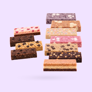 A set of assorted protein bars and nourish bites arranged in two groups against a solid light purple background. The bars feature various toppings and fillings, including chocolate chips, dried fruit, nuts, and white chocolate pieces, in shades of brown, beige, pink, and purple.