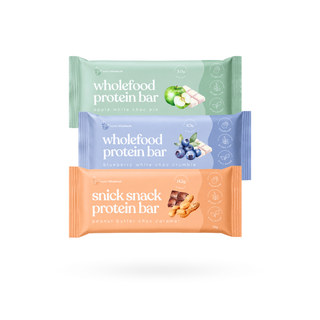Three packaged protein bars stacked vertically on a white background: a  green Wholefood Protein Bar (apple white chocolate pie), a lavender-blue Wholefood Protein Bar (blueberries white chocolate crumble), and an orange Snick Snack Protein Bar (peanut butter choc caramel).