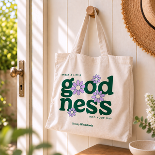A natural tote bag hanging on a wooden door hook in a bright, sunlit entryway. The bag features a retro-style graphic with the words 'goodness' in dark green, accented with purple and white flowers, and the phrases 'SNEAK A LITTLE goodness INTO YOUR DAY' and 'Sneaky Wholefoods'.