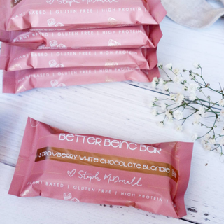 Pink packaging of Better Being Bar strawberry white chocolate blondie on a white surface with flowers.