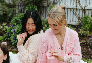 Three women in pink cardigans sitting outdoors on a blanket with flowers.
