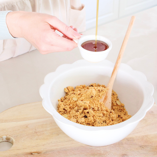Person pouring maple syrup into a bowl of protein ball mix on a wooden surface.