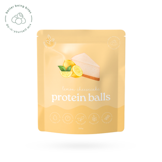 A stand-up pouch package for lemon cheesecake protein balls, shown on a plain white background. The pouch is a warm pale yellow-orange with an illustration of a slice of cheesecake and lemons on the front, plus the text 'lemon cheesecake protein balls' and small icons along the bottom.