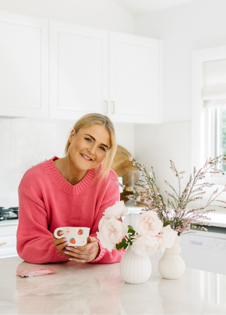 Woman in a pink sweater holding a mug in a bright kitchen with flowers on the counter.