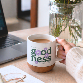 Person holding a mug with 'goodness' text, sitting at a desk with a laptop and vase.