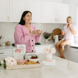two friends enjoying Sneaky Wholefoods Strawberries and Cream Protein Bars during a relaxed morning tea, paired with fresh juice and coffee, embodying a delightful way to stay nourished and connected